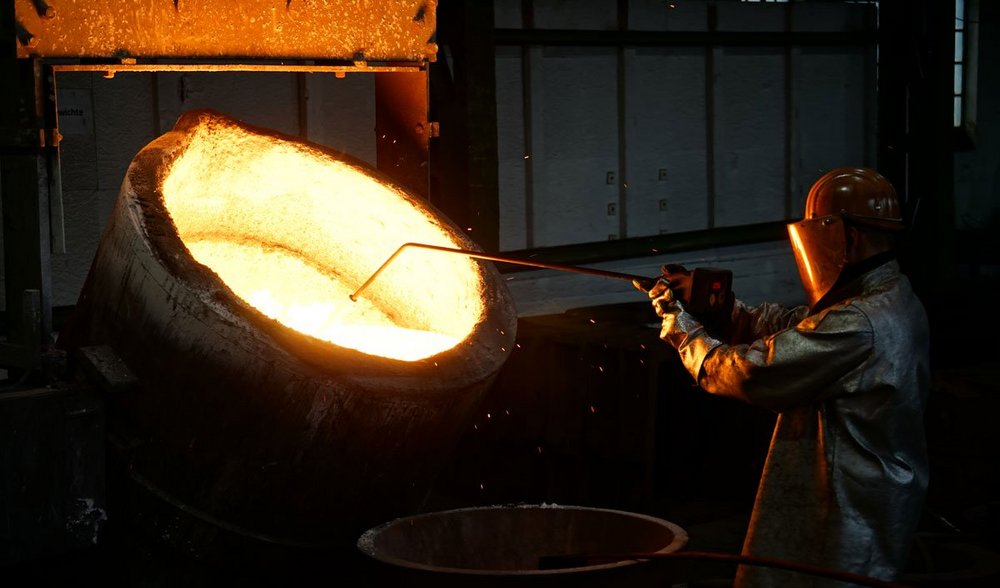 Foundry worker in protective gear skimming impurities from molten metal in a large ladle at a foundry.