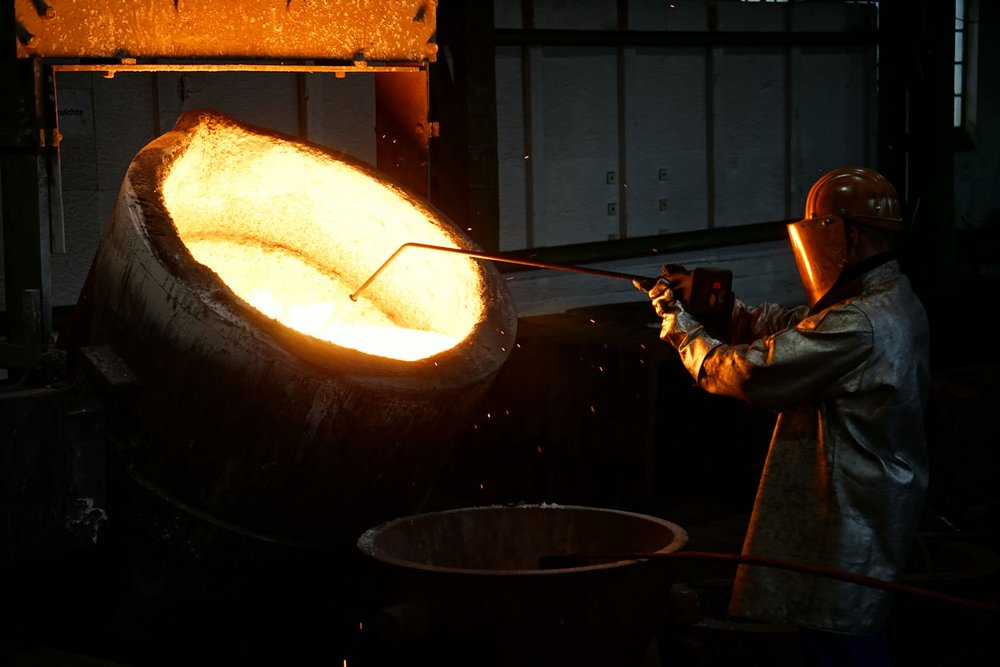 Foundry worker in protective gear skimming impurities from molten metal in a large ladle at a foundry.