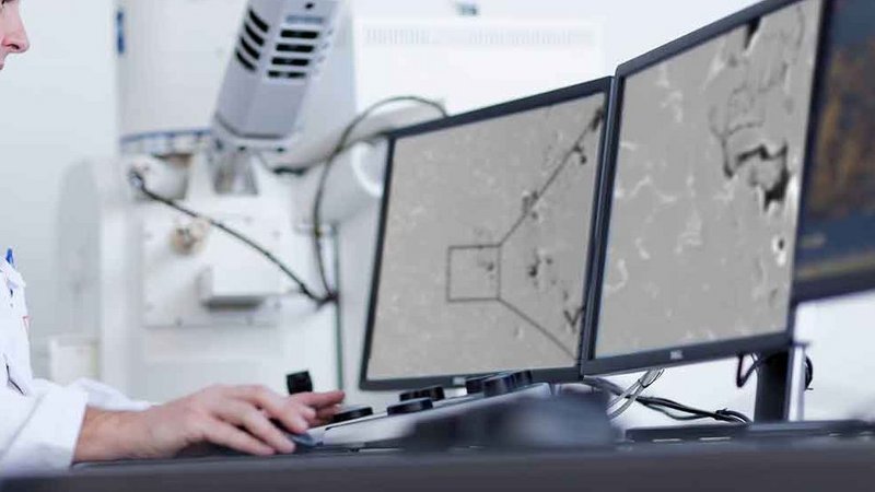 Scientist analyzing samples with a scanning electron microscope, displayed on multiple monitors in a laboratory setting.