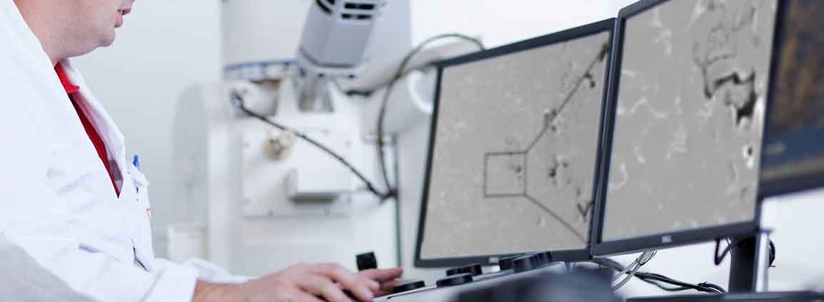 Scientist analyzing samples with a scanning electron microscope, displayed on multiple monitors in a laboratory setting.