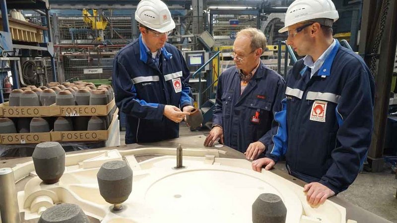 Two technicians demonstrating a feeder sleeve to a foundry worker, with a mold and additional Foseco feeder sleeves in the background