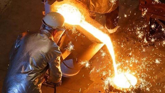 Overhead view of a foundry worker in protective gear pouring molten metal from a furnace into a mold, with bright sparks flying around the intense and fiery work environment.