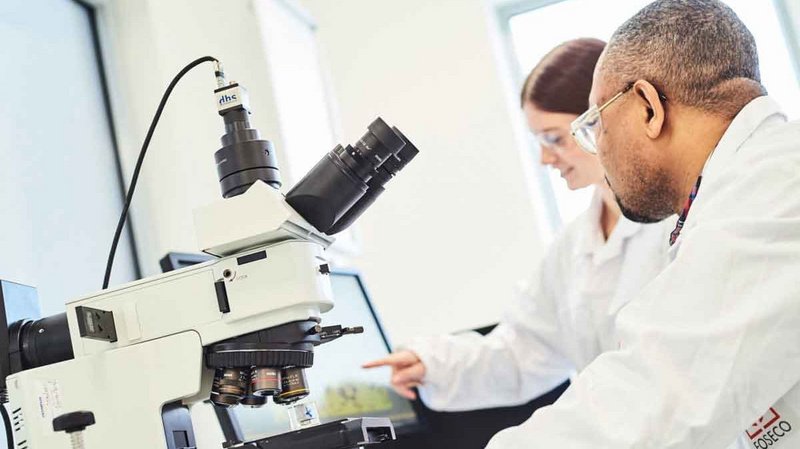 Two laboratory technicians in white coats examining metal casting samples under a microscope to assess quality in a bright laboratory setting