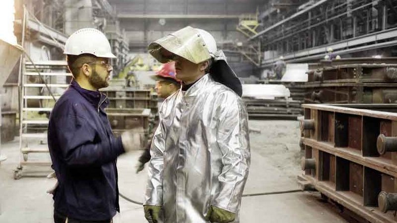  Two foundrymen engaged in discussion, wearing protective helmets and clothing, with a large ladle pouring molten metal in the background of a busy foundry