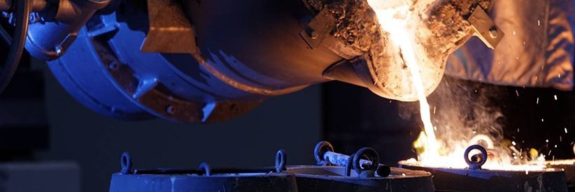 Wide-angle view of a large foundry crucible pouring glowing molten metal into molds, with sparks and flames against a dark industrial background.