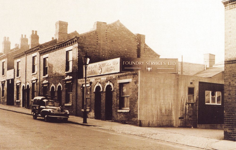 Historical sepia-toned photo showing Foundry Services Ltd storefront on a quiet street with vintage cars parked and traditional brick houses in the background