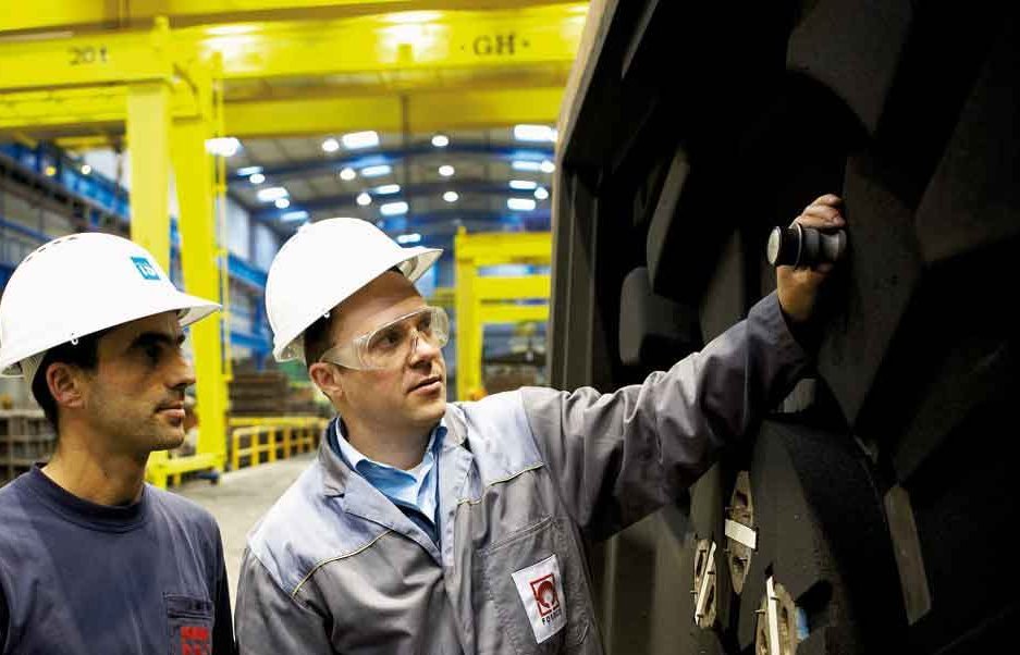 A Foseco specialist explains the properties of mold binders to a foundry worker beside large industrial machinery in a manufacturing setting