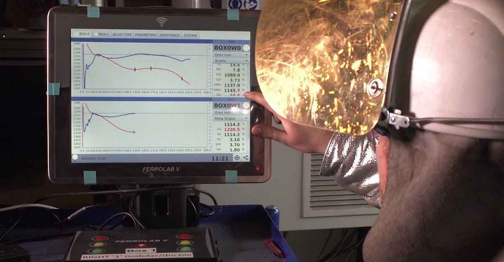 Technician monitoring metal temperature on a FERROLAB V device screen with molten metal in the background, wearing safety gear.